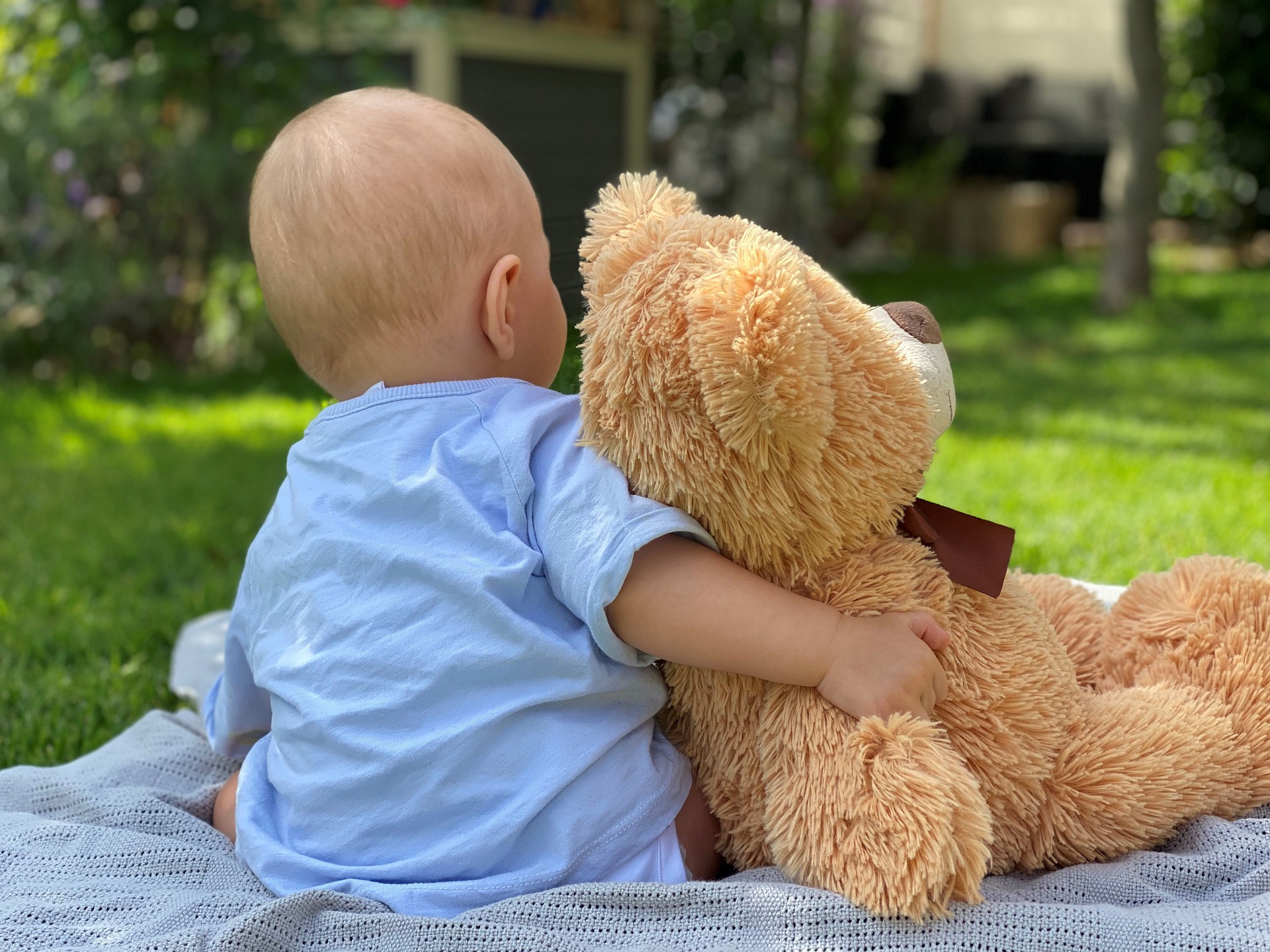 Kuvassa vauva, jolla on vaaleansininen paita, istuu selin kuvaajaan päin viltin päällä nurmikolla ja hänellä on ruskea, iso nalle vieressään kainalossa.