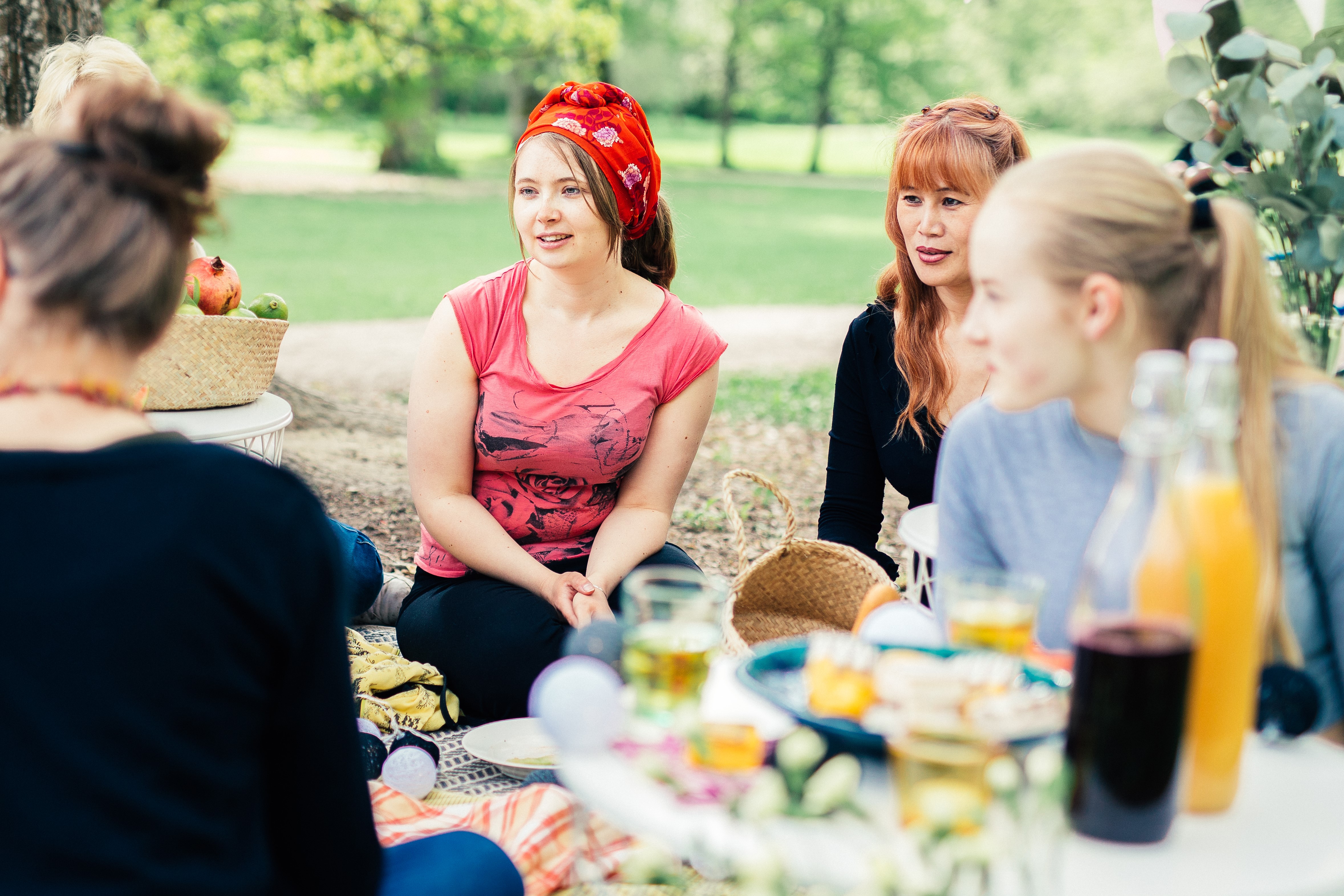 A group of young adults sits on a blanket in the park with picnic snacks in front of them.