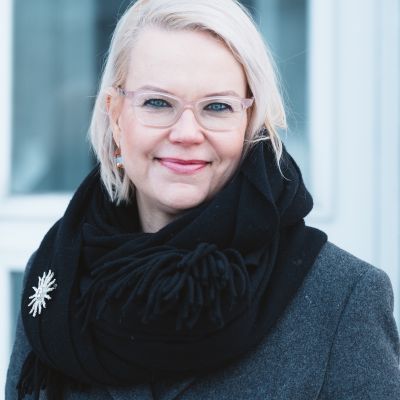 Vaaleahiuksinen Soile hymyilee vaaleasankaisissa silmälaseissa kohti kameraa kaulassa musta huivi.
