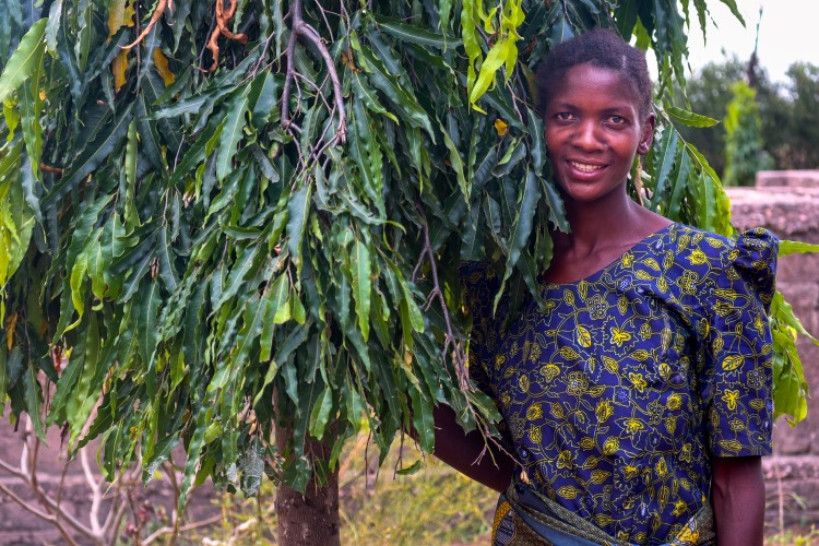 A smiling woman stands next to a broad-leafed tree.
