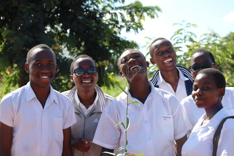 A group of smiling Tanzanian youths with a tree sapling in front of them.