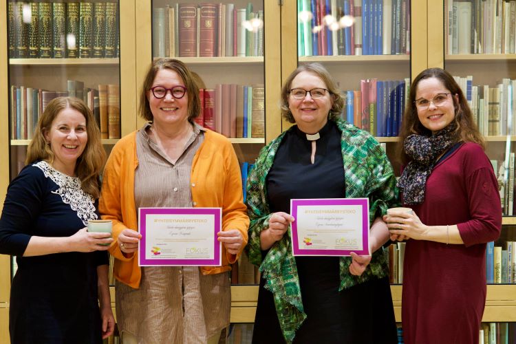 Neljä naista hymyilee kameralle kirjahyllyn edessä, kahdella heistä on diplomi kädessä.