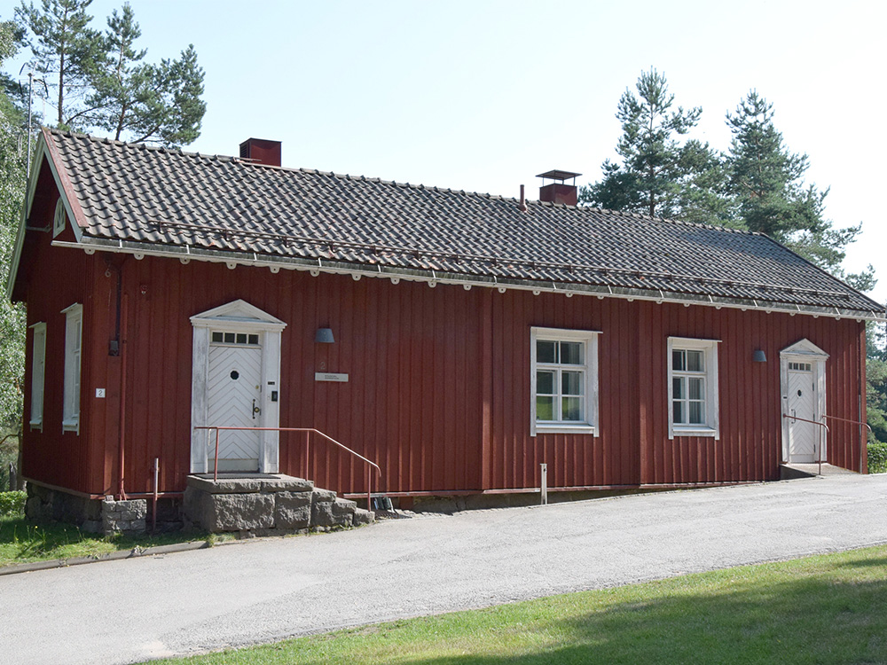 Punamullattu vanha pitkä hirsitalo, jossa on valkoiset koristeelliset ovat, punainen tiilikatto ja valkokehyksiset ruutuikkunat.