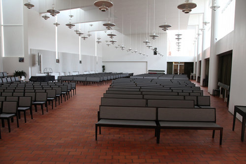 Sisäkuva kirkkosalista. Kuvassa riippuvia kattovalaisimia, tuoleja ja penkkejä.