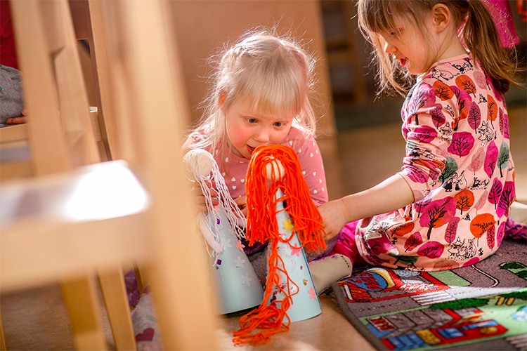 Kaksi pientä tyttöä leikkii keskittyneesti itse tekemillään nukeilla.