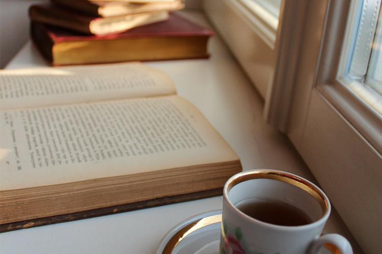 Kirjojaa pinossa ikkunalaudalla. Edessä kultareunuksinen kuppi teetä.