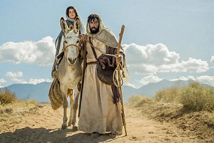 Aavikolla, vuorten ja pilvien edessä mies (Joosef) seisoo aasin vieressä, aasin selässä istuu nainen (Maria)