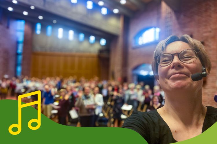Kuva kirkkosalista. Etualalla kuoron ohjaaja, taustalla paljon osallistujia kirkkosalissa. Kuva otettu selfienä.