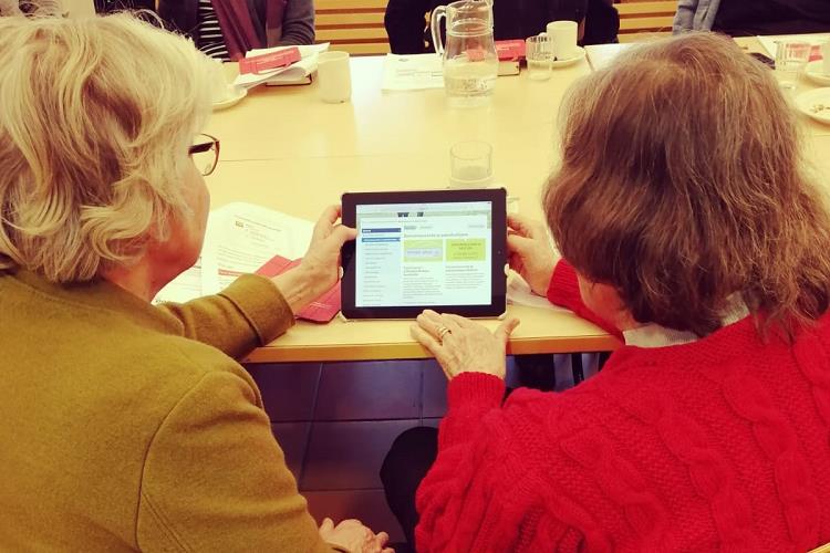 Kuvassa kaksi naista istuvat vierekkäin pöydän ääressä, selkä kameraan kohti, Pitävät yhdessä käsissä tablettia.