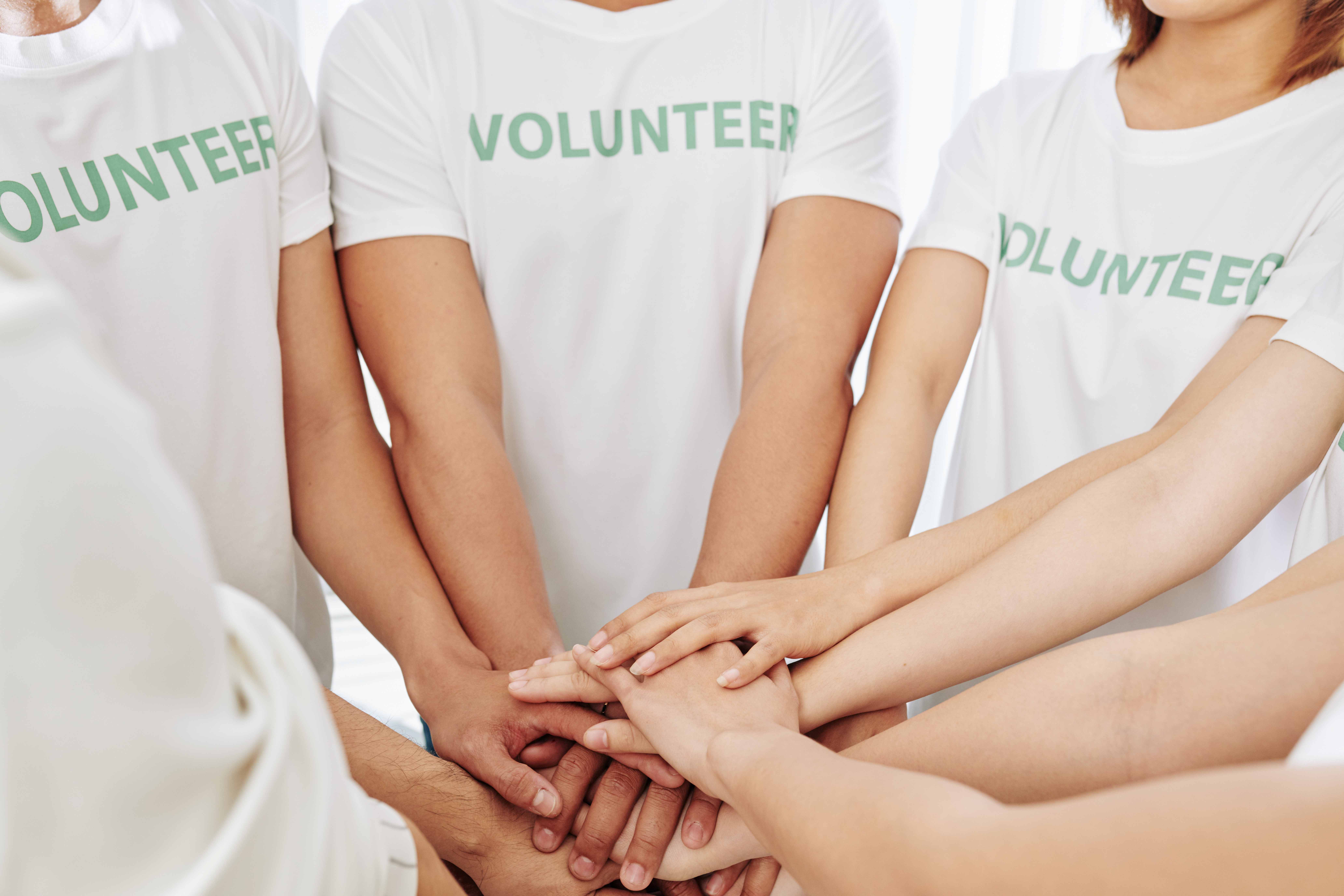 Kuvassa on henkilöitä, joiden valkoisessa t-paidassa lukee teksti: volunteer. He ovat asettaneet kätensä yhteen. 