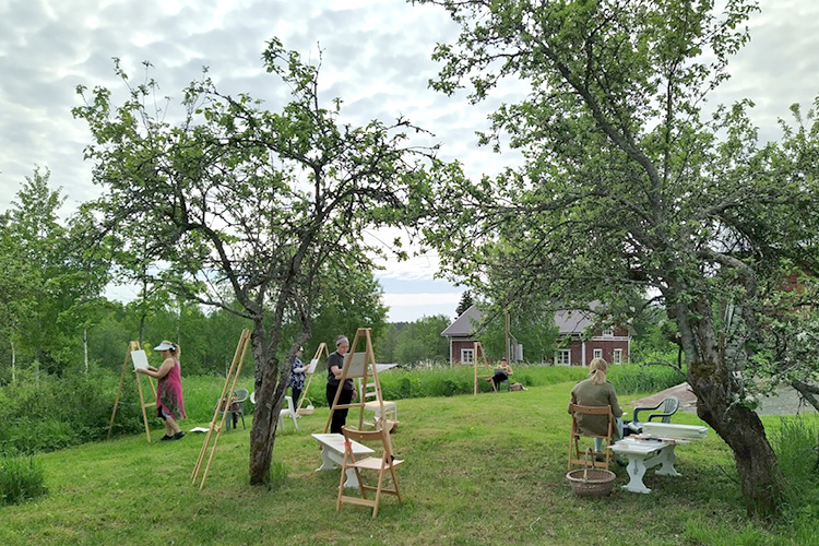 ihmisiä maalaamassa tunnelmallisessa puutarhassa omenapuiden alla, taivas on poutapilvessä