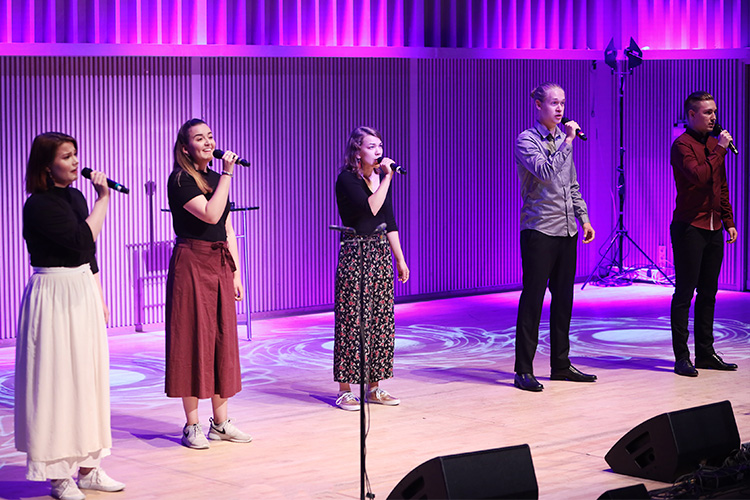 kolme naista ja kaksi miestä laulavat mikrofoneihin, valon väri on violetti