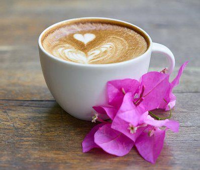 Valkoisessa kahvikupissa kahvi sydänkoristeella, liila kukka kupin vierellä.