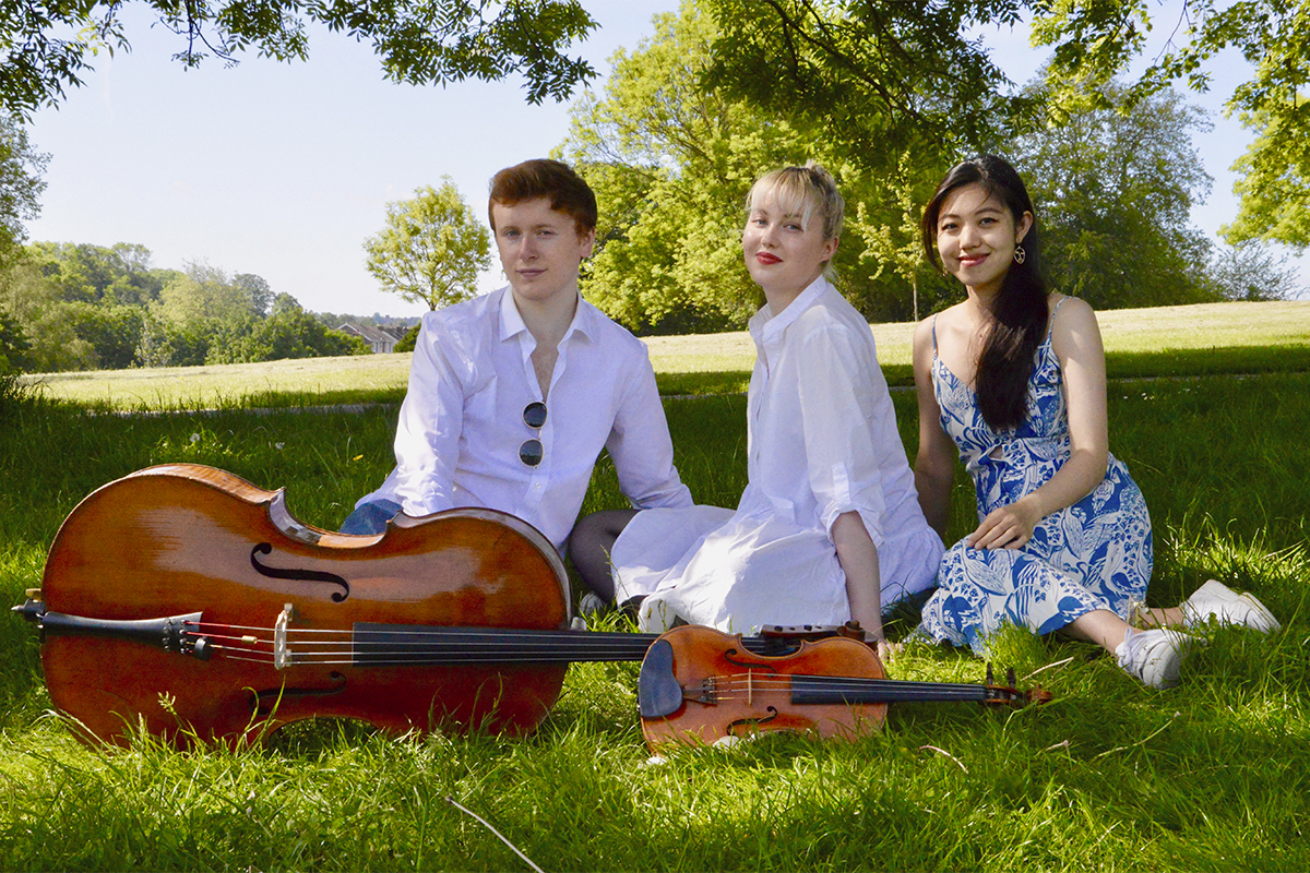 Kuvassa kolme nuorta muusikkoa istuu nurmikolla. Heillä on kuvassa mukanaan myös sello ja viulu.