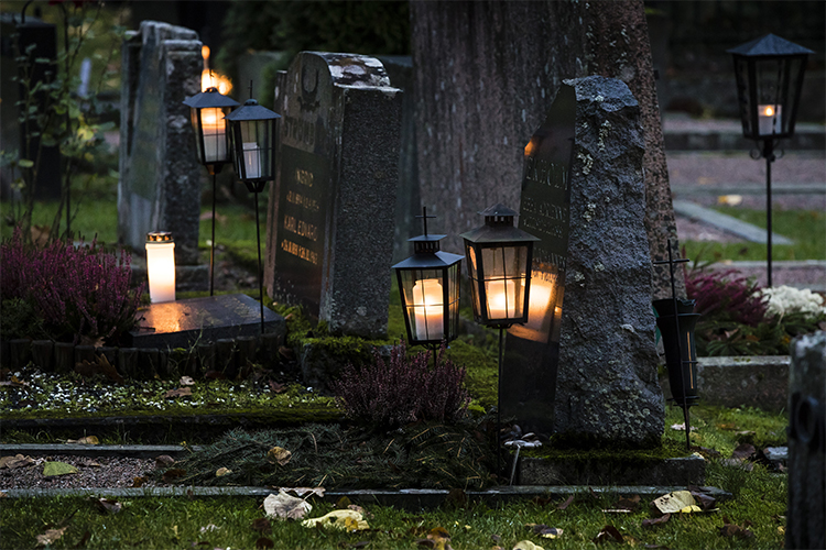 Kuvassa on kynttilälyhtyjä hautakivien edessä illan hämärässä.