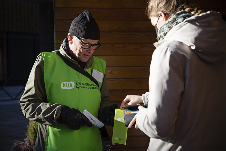 Kuvassa on kaksi ihmistä, joista toisella on vihreä keräysliivi päällään ja lipas kädessään, johon toinen l...