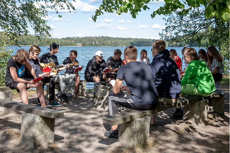 Kuvassa ryhmä nuoria istuu laulamssa puupenkeillä järven rannassa.