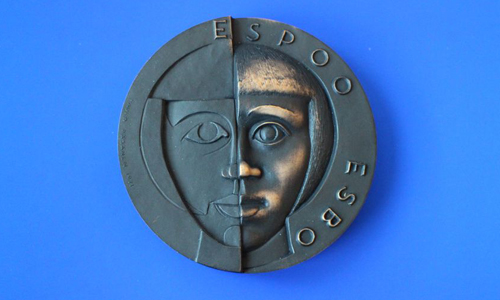 Espoo-mitalin etupuolella on tyylitellyt kasvot ja takana puoli-istuva hahmo.