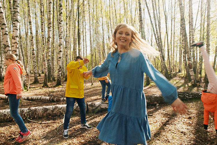 nainen tanssii etualalla sinisessä mekossa, taustalla lapset tanssivat, takana keväinen koivikko