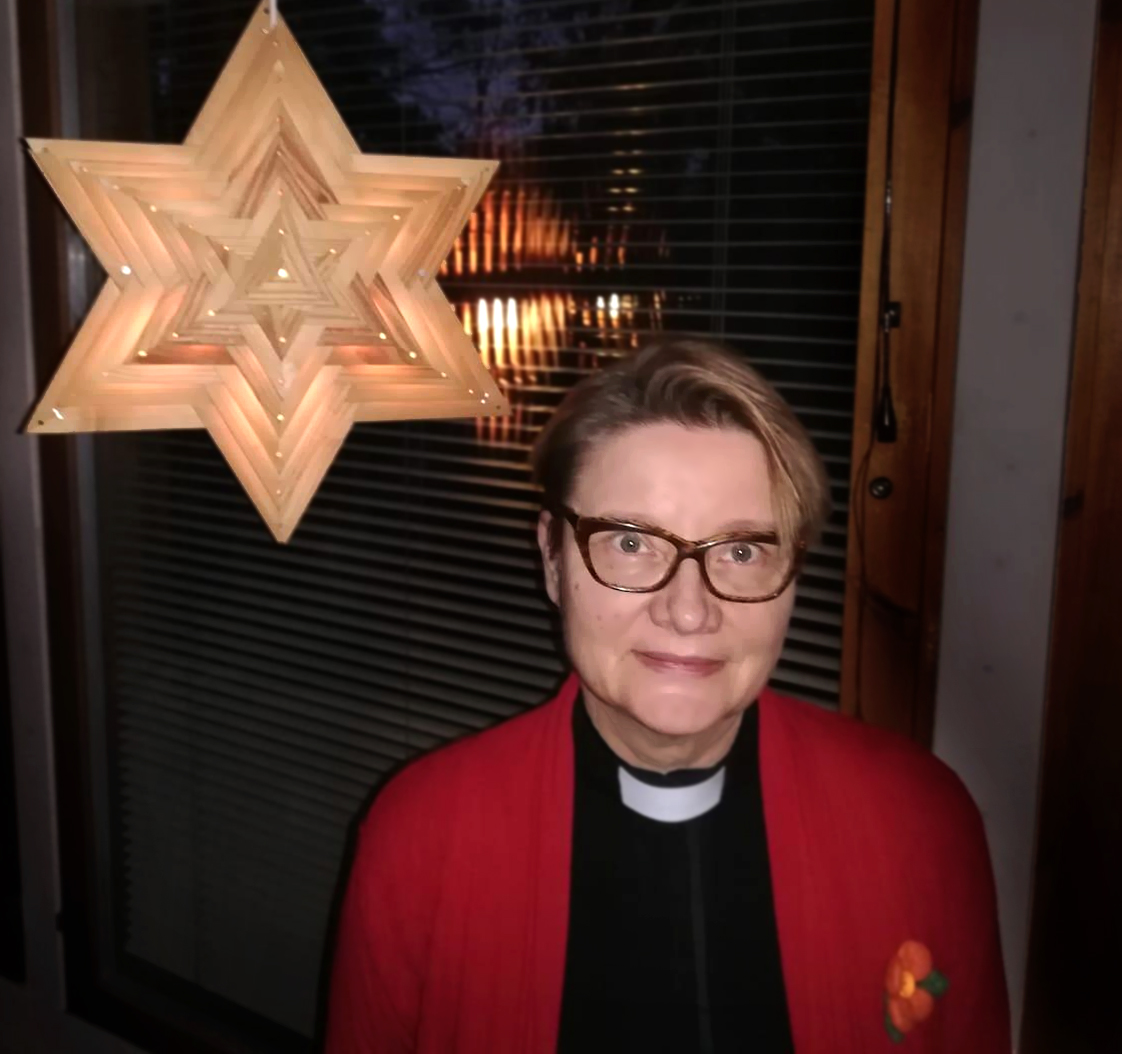 Kuvassa on kirkkoherra Päivi Linnoinen ikkunan edessä, vieressään joulutähti, jossa on valot.