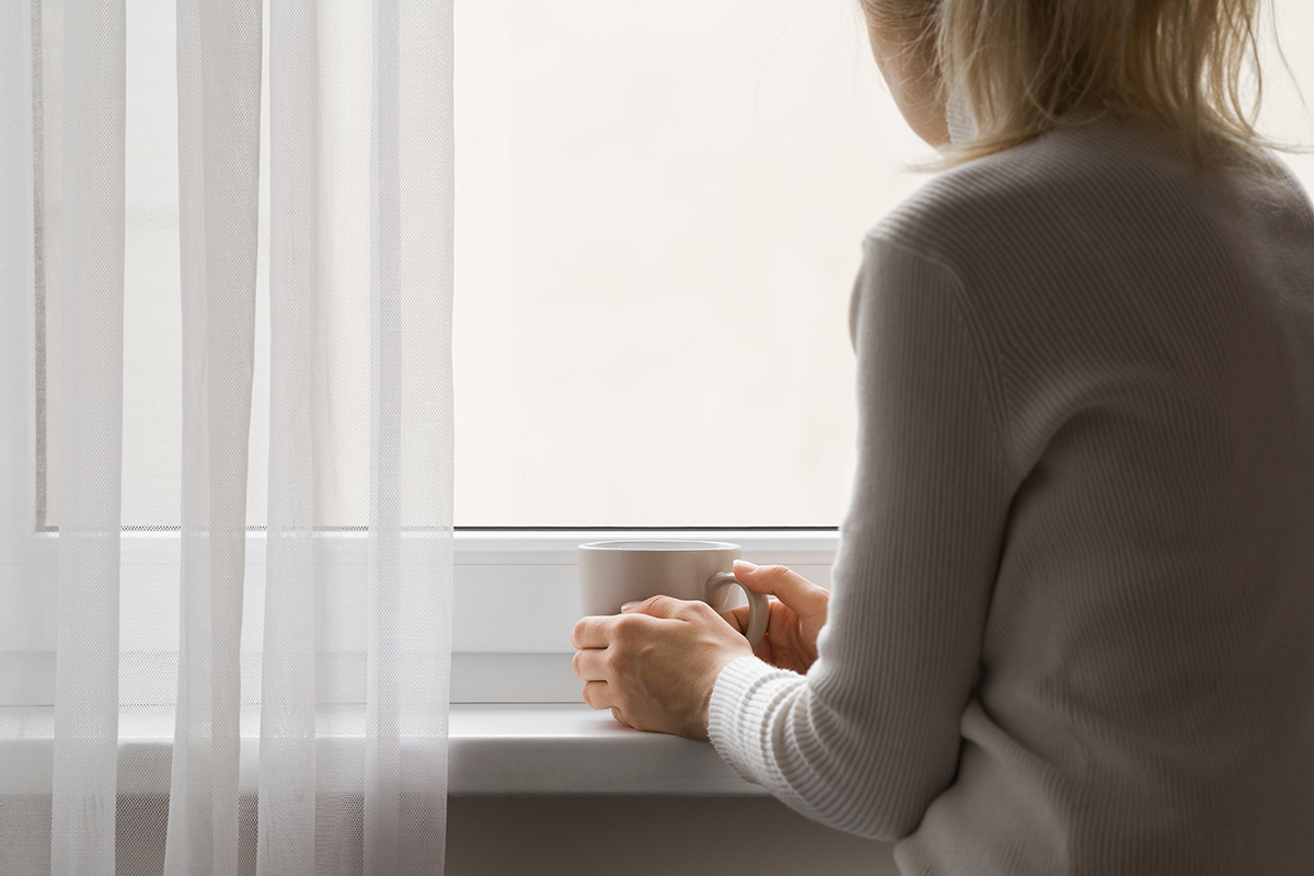 Kuvassa yksinäinen nainen katsoo ulos ikkunasta. Käsissä on kahvimuki.