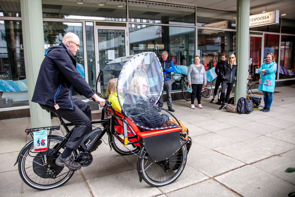 Kuvassa ajetaan riksapyörällä Tapiolan keskustassa, pyörän kyydissä on kaksi ihmistä.