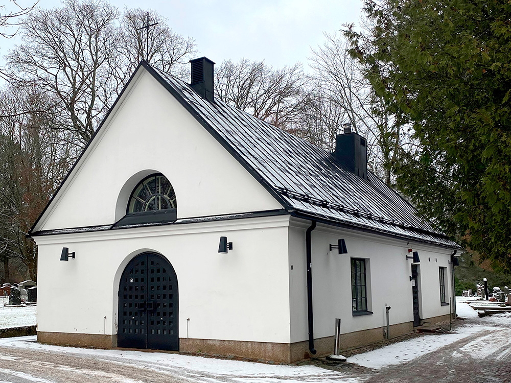 Valkoinen mustakattoinen rakennus, harjakatto, kaariovi ja -ikkuna.