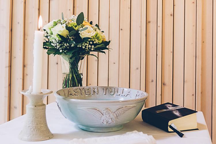 Kastepöydällä palaa kynttilä, on kukkakimppu, kastemalja ja raamattu.