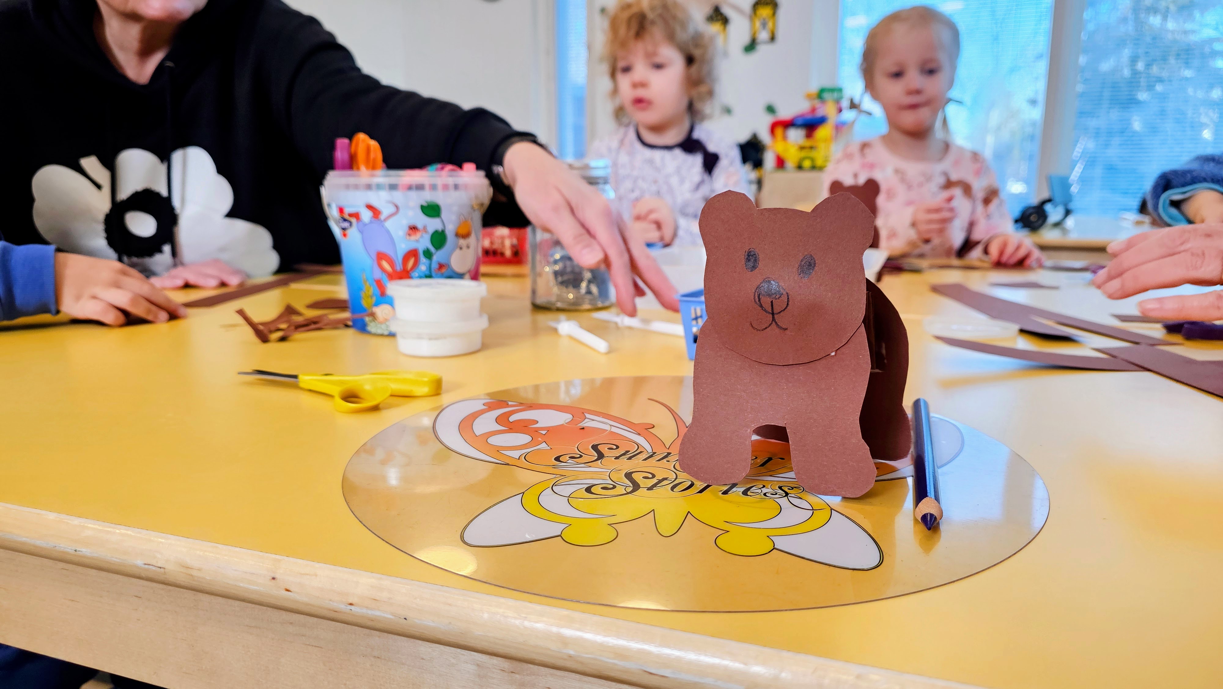 Kartongista leikattu karhu ja askartelutarvikkeita pöydällä, jonka ääressä lapsia ja aikuisia.