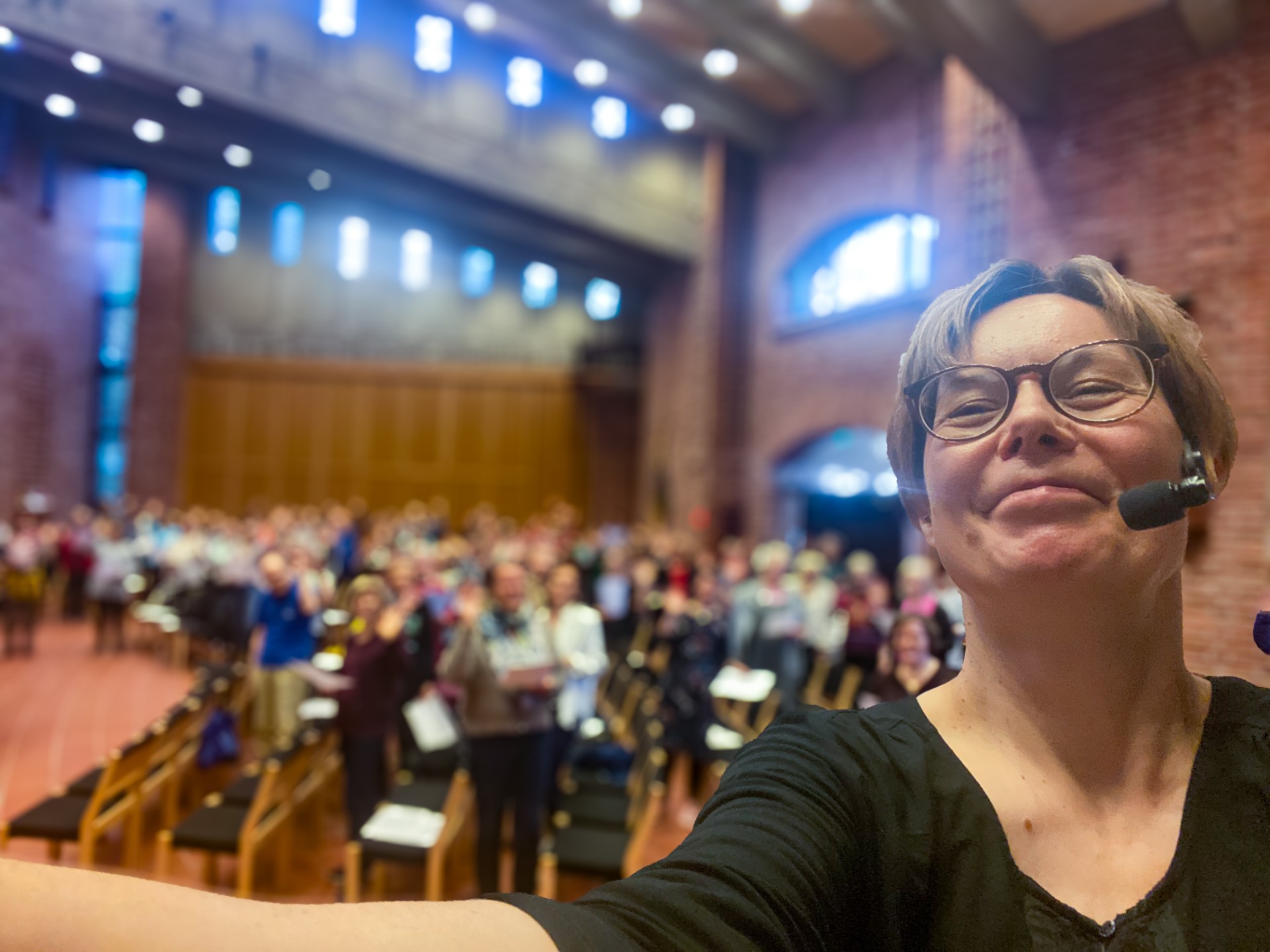 Selfiekuva kirkkosalista, jossa kuorolaisia harjoituksia sali täynnä. Etualalla kuoro ohjaaja Outi.