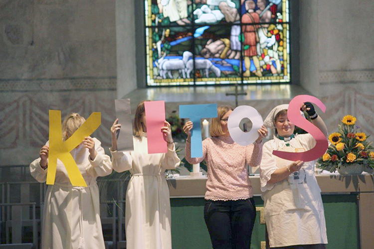 Viisi naista pitää kirkon alttarilla isoja kylttejä, joista muodostuu sana "kiitos".